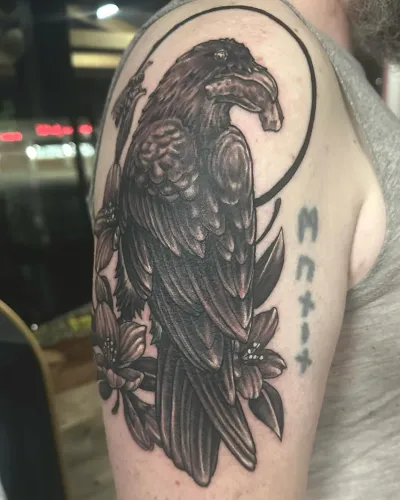 Black and grey raven perched below a full moon on an upper arm — by Adam Crane at Ministry Ink