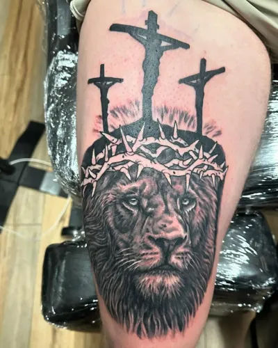 Black and grey lion wearing a crown of thorns beneath three crosses, on a thigh — by Adam Crane at Ministry Ink
