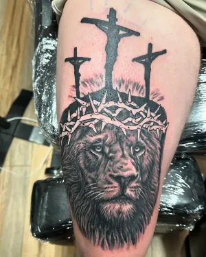 Black and grey lion wearing a crown of thorns beneath three crosses, on a thigh — by Adam Crane