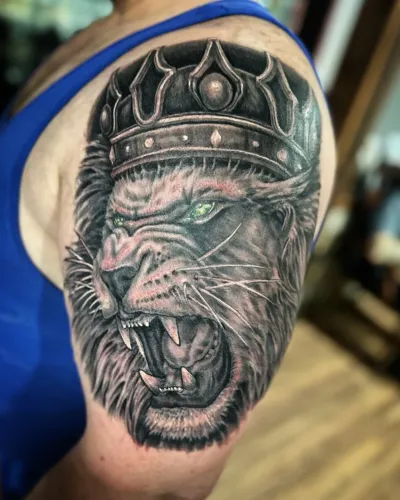 Black and grey roaring lion wearing a jewelled crown on a shoulder — by Adam Crane at Ministry Ink