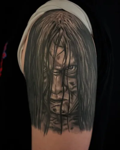 Black and grey portrait of a long-haired horror figure on an upper arm — by Adam Crane at Ministry Ink