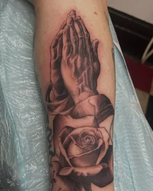 Black and grey praying hands with a rose on a forearm — by Gabriel Gonzalez