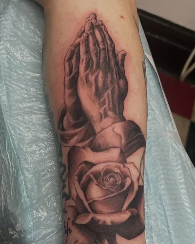 Black and grey praying hands with a rose on a forearm — by Gabriel Gonzalez at Ministry Ink